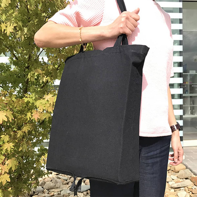 A black foldable calico tote bag held by a person against a backdrop of greenery and modern architecture.