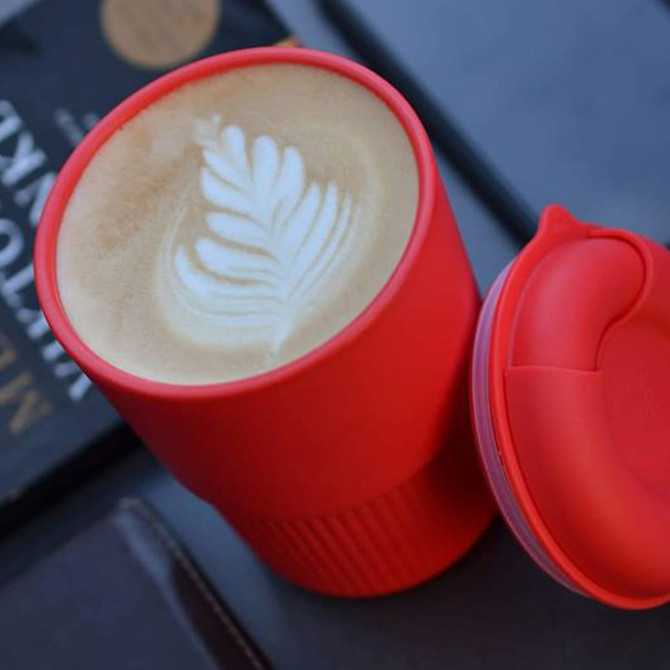 A reusable coffee cup in vibrant red with a latte art design on the frothy surface, featuring a lid and a textured grip.