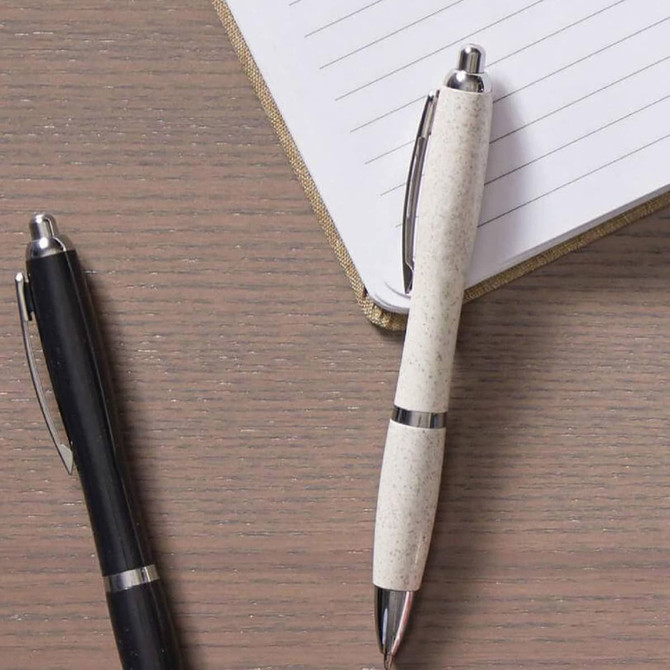 A black pen and a white eco-friendly pen sit on a wooden surface next to a lined notebook. Both have metal accents.