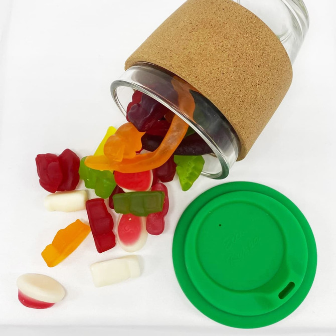 A glass coffee cup with a cork band, filled with colourful jelly lollies, and a green lid beside it.