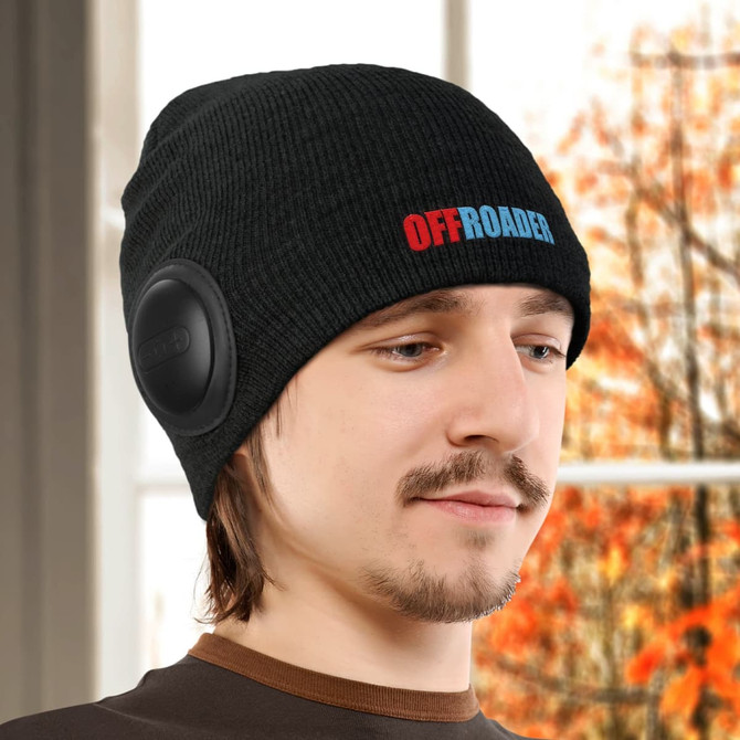 A black beanie with built-in speakers, featuring a logo and worn by a person in a warm, indoor setting.