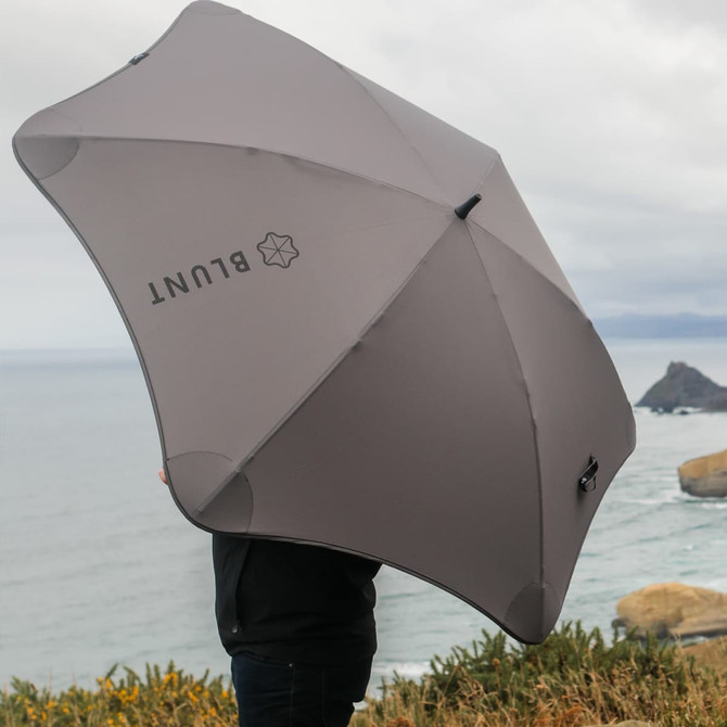 A grey BLUNT Sport Umbrella held by a person against a coastal backdrop with cloudy skies.