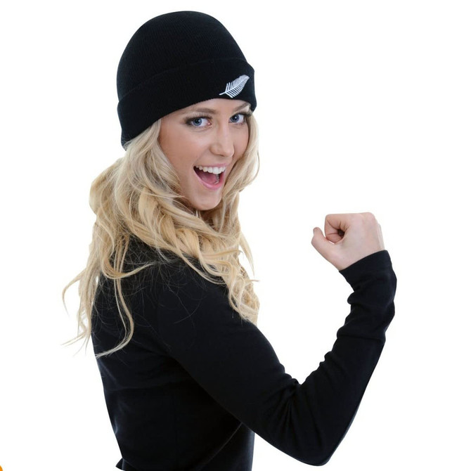 A black beanie with a silver fern logo, worn by a smiling person flexing their arm.