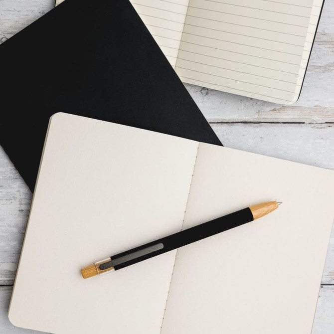 A black aluminium pen with a wooden accent rests on an open blank notebook, alongside a black notebook.