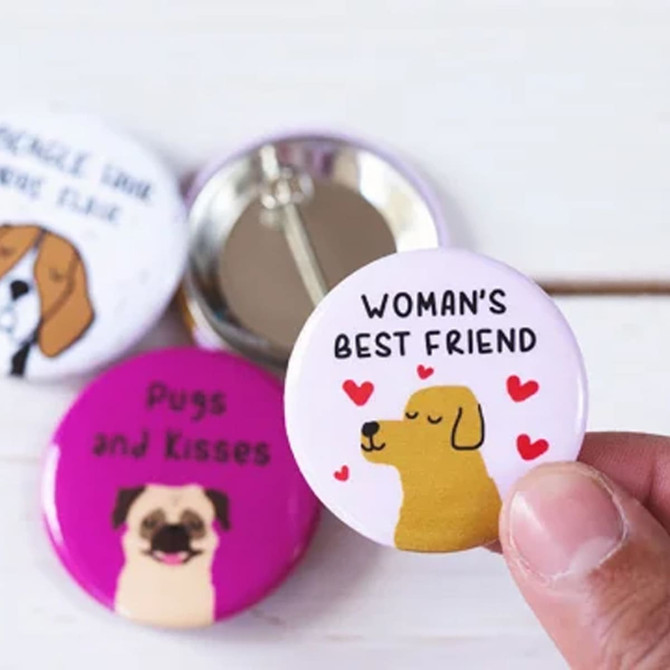 A hand holds a round, 25mm button badge featuring a dog illustration on a pink background, among other badges.