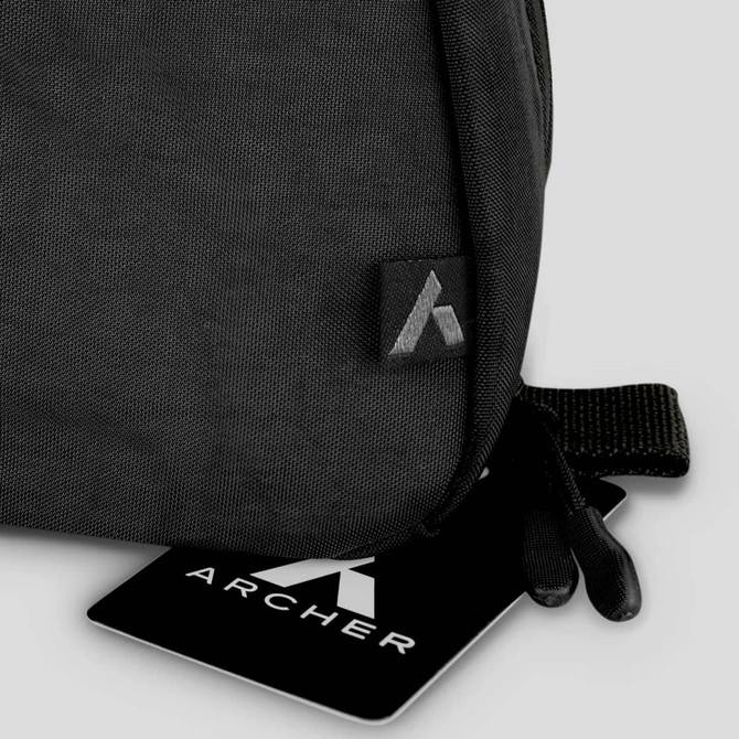 A black toiletry bag with a textured fabric and a small logo. It features a zip closure and a hanging loop.