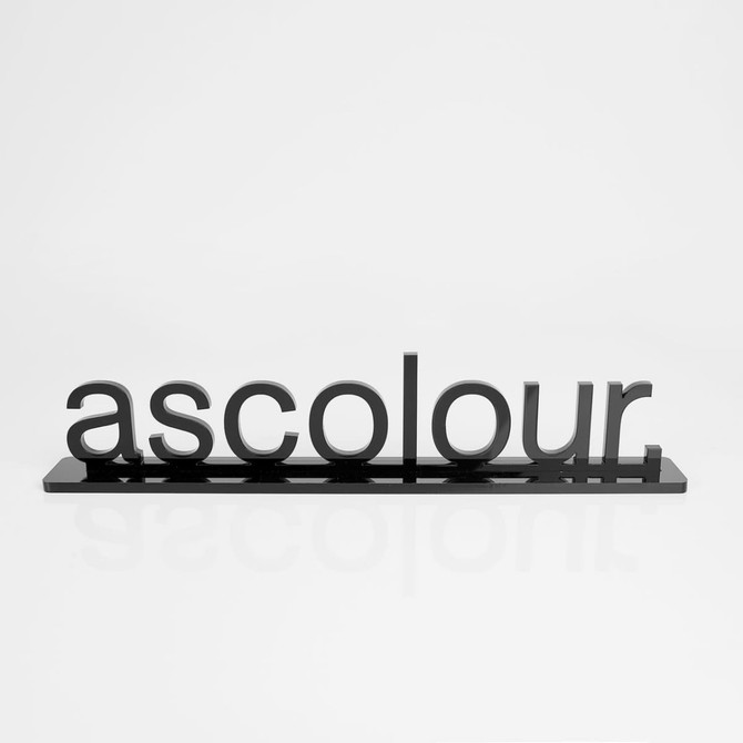 A black sign displaying the word "ascolour" in three-dimensional letters on a reflective base.