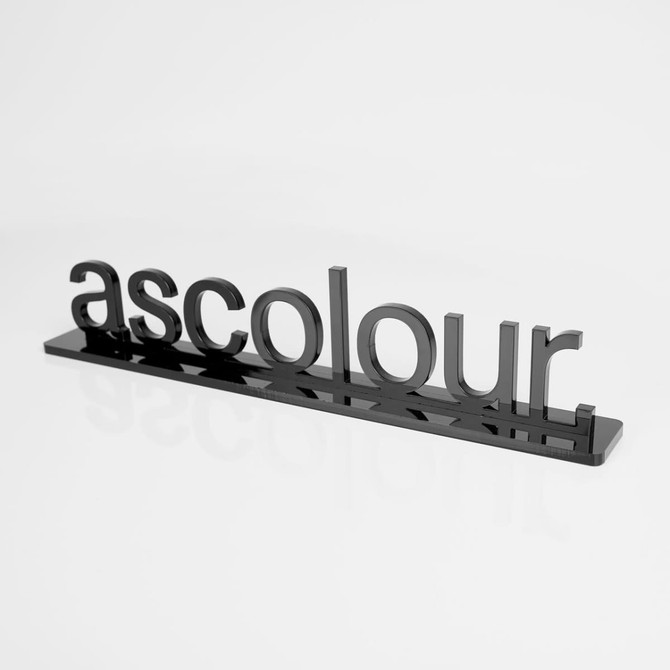 A black acrylic display sign featuring the word "ascolour" in bold letters, set against a white background.