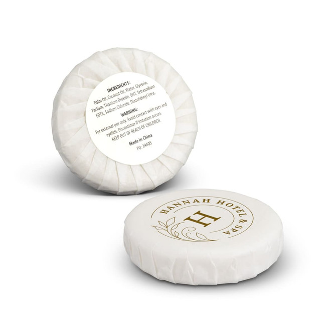 A round white soap bar, wrapped elegantly, featuring a logo and gold detailing, with listed ingredients on the reverse side.