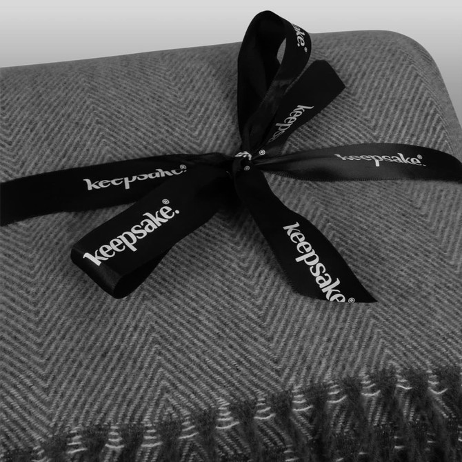A grey throw blanket tied with a black ribbon featuring a logo, displaying a textured pattern and fringe edges.