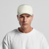 A cap in a light cream colour, featuring a structured design and a logo on the side, worn by a model.
