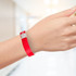 A red plastic wristband is displayed on a hand, featuring a silver fastening. The background is blurred and light-filled.