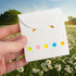 A hand holds a white envelope with colourful sticky note pads peeking out, surrounded by a field of daisies.