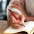 A metal pen in vibrant red held by a hand above an open notebook, featuring a logo on the side.