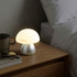 A mushroom-shaped table lamp in soft white, with a silver base, sits on a bedside table next to glasses and a mug.