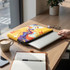 A vibrant yellow laptop sleeve with abstract designs placed on a wooden table beside a coffee cup and notebook.