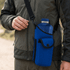 A drink bottle nestled in a blue neoprene bottle holder, featuring a strap and pocket, attached to a person's side.