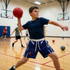 A boy wearing navy blue active shorts and a black t-shirt throws a red dodgeball in a gym setting.