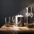 A glass carafe with ice, accompanied by three dark grey glasses and a silver ice bucket, all set on a wooden surface.