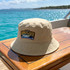A beige bucket hat with a logo sits on a wooden table beside a turquoise sea under a clear sky.