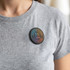 A round, multi-coloured magnetic badge worn on a grey shirt, featuring a logo and text design.