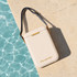 A cream cooler bag with a grey strap, featuring a logo, resting on a stone surface by water.