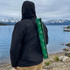 A green cooler sling bag is carried on the back of a person by the water, with mountains in the background.