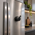 A compact black speaker adheres to a stainless steel refrigerator in a modern kitchen setting.