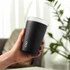 A drink bottle in matte black with a translucent top and a logo. It is held in a hand against a blurred background.