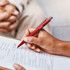 A red plastic pen held in a hand, with a clipboard featuring a checklist in the background. The pen has a logo.