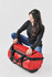 A medium red waterproof gear bag with black straps and a logo, held by a person seated on the floor.