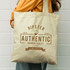 A cream tote bag with a large branding logo, featuring brown text and a casual design.