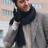 A man wearing a black scarf and black gloves, holding a phone, dressed in a grey patterned jacket.
