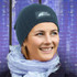 A blue cable knit beanie with a patch, worn by a smiling person with a light scarf, against a purple background.