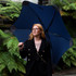 A navy umbrella with a unique shape is held by a person in a black blazer, surrounded by greenery.