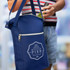 A navy blue cooler bag with a logo, featuring a zip pocket and carrying straps, holding a drink bottle.