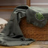 A green herringbone throw blanket draped over a woven basket, featuring a small logo.