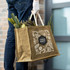 A jute tote bag in brown, featuring a logo and filled with fresh vegetables, held by a person wearing gloves.