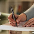 A black pen is held by a hand, writing on a white sheet of paper, with a blurred background.
