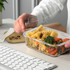 A clear glass lunch box filled with pasta, vegetables, and a fork, beside a keyboard and a wooden lid.