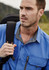 A man wearing a blue short sleeve button-up outdoor shirt, carrying a backpack, with a scenic background.