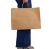 A large brown paper bag with handles, held by a person wearing blue jeans and beige shoes.