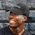 A black cap with a logo, featuring a curved brim, worn by a smiling individual against a textured stone background.