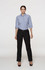 A woman stands in black pants and a blue checkered shirt, featuring rolled sleeves and a belt. The image includes branding.