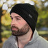 A black beanie featuring a textured knit design and a logo, worn by a man with a beard, set against a blurred outdoor background.