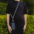 A phone lanyard in white is attached to a smartphone, worn by a person standing in front of greenery.