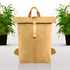 A yellow kraft paper backpack with a roll-top closure and adjustable straps, placed among green potted plants.
