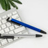 A blue stylus pen and a silver plastic pen rest on a grey keyboard, with a green leaf in the background.
