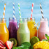 Colourful paper drinking straws in jars filled with various smoothies, surrounded by fresh fruit and greenery.