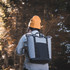 A grey cooler totepack is worn on the back of a person in a black and orange beanie, surrounded by trees.